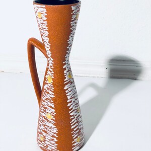 Puede incluir: Un jarr&oacute;n de cer&aacute;mica marr&oacute;n con un patr&oacute;n geom&eacute;trico blanco y amarillo. El jarr&oacute;n tiene un asa y una abertura estrecha.