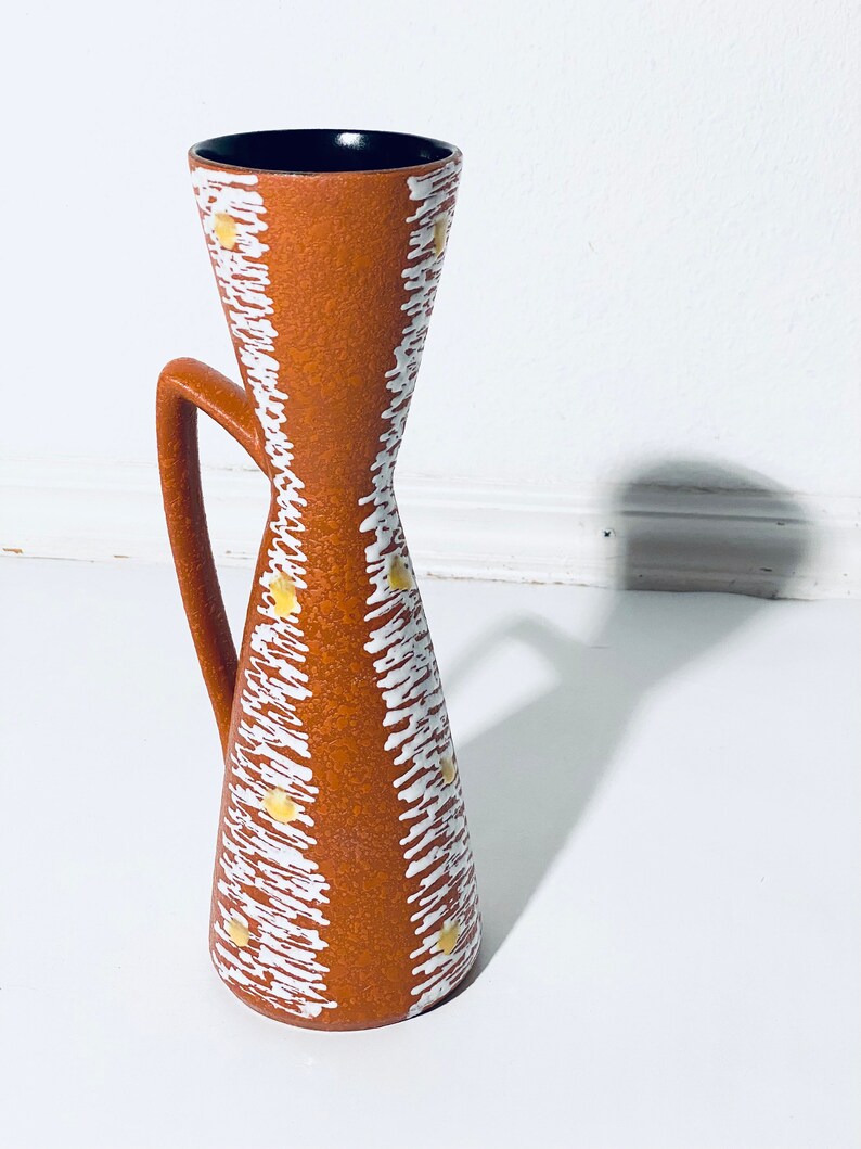 Puede incluir: Un jarr&oacute;n de cer&aacute;mica marr&oacute;n con un patr&oacute;n geom&eacute;trico blanco y amarillo. El jarr&oacute;n tiene un asa y una abertura estrecha.