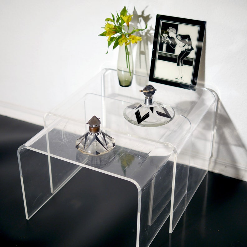 May include: Two clear acrylic tiered tables with a black and white art print, a yellow flower arrangement in a clear glass vase, and two decorative glass bottles with black and white geometric designs.