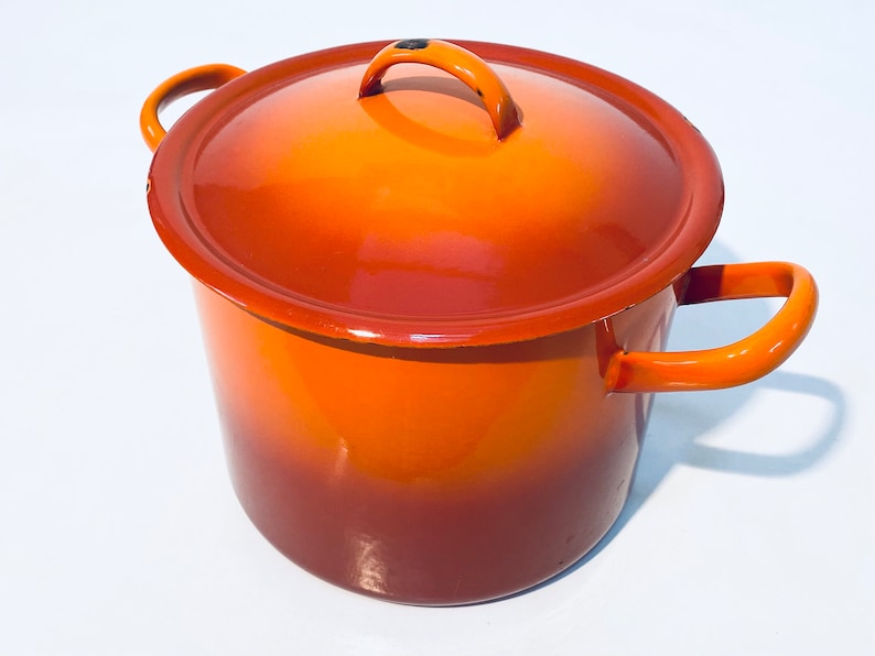 Pu&ograve; includere: Pentola smaltata arancione con coperchio e due manici. La pentola ha un gradiente di colore dall'arancione al marrone.