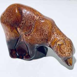 Può includere: Una statuetta di orso in ceramica marrone con una finitura testurizzata. L'orso è in posizione accovacciata con la testa bassa, come se stesse annusando il terreno.