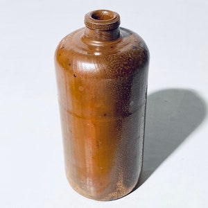 Könnte beinhalten: Eine braune Keramikflasche mit einer breiten Öffnung und einer leicht abgerundeten Form. Die Flasche hat eine strukturierte Oberfläche und eine hellbraune Farbe.