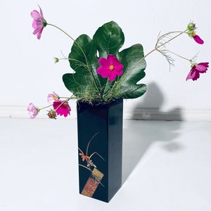 Könnte beinhalten: Eine schwarze Vase mit goldenen Akzenten und einem Blumendesign enthält rosa und magentafarbene Kosmeen und ein großes grünes Blatt.