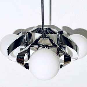 May include: A modern chrome and white pendant light fixture. The light has a series of curved, polished metal bands that surround six spherical, frosted glass globes. The fixture is suspended from a single chrome rod.