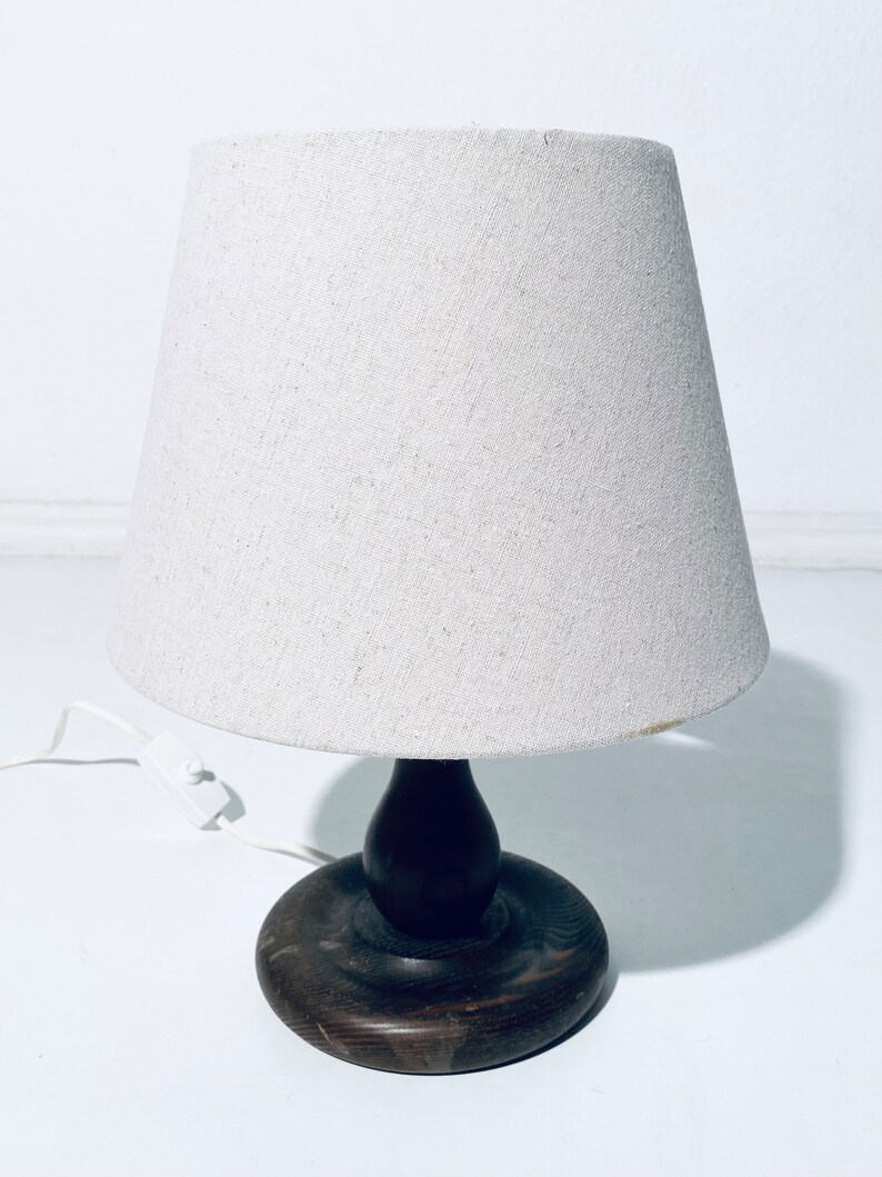 May include: A wooden table lamp with a white fabric lampshade. The lamp has a dark brown wooden base with a round, flat top and a bulbous, rounded stem.