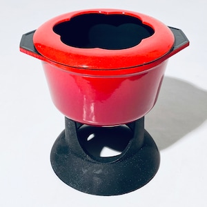 Könnte beinhalten: Rote Fondue-Pfanne mit schwarzem Sockel und einer blütenförmigen Öffnung im Deckel.