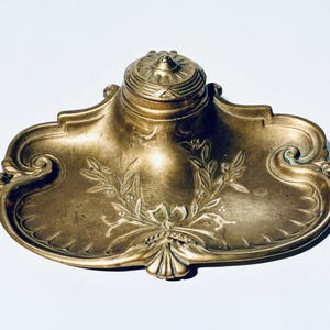 Könnte beinhalten: Verzierter antiker Tintenfass aus Messing mit dekorativem Blumen- und Blattdesign. Das Tintenfass hat einen zentralen Tintenbehälter mit Deckel und eine Tablettbasis mit gerollten Kanten. Ein klassisches Schreibtischzubehör.