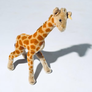 Könnte beinhalten: Ein Plüsch-Giraffen-Spielzeug mit langem Hals und Fleckenmuster in Braun- und Beigetönen. Die Giraffe hat kleine Ohren und ein detailliertes Gesicht und steht auf vier Beinen. Das Spielzeug besteht aus weichem, strukturiertem Material.