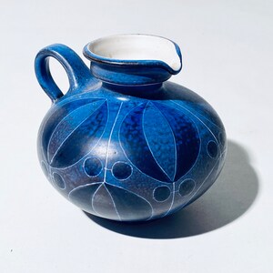 Könnte beinhalten: Eine blaue Keramikkanne mit weißem Innenraum und geometrischem Muster. Die Kanne hat einen runden Körper und einen kleinen Ausguss.
