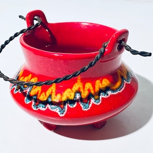 Könnte beinhalten: Ein roter Keramikkessel mit einem schwarzen Drahtgriff und einem gelben und blauen Flammenmuster. Der Kessel hat drei kleine Füße.