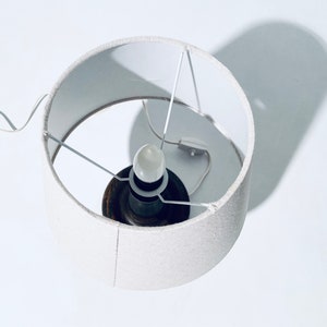 May include: A white lampshade with a white cord and a light bulb inside. The lampshade is made of a textured fabric.