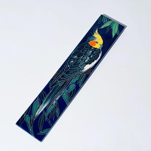 Könnte beinhalten: Rechteckige dekorative Fliese mit einer detaillierten Illustration eines Nymphensittichs. Der Vogel hat gelbe und orangefarbene Kopf federn, mit einem dunkelblauen und grünen Körper. Der Hintergrund ist dunkelblau mit grünen Blättern.