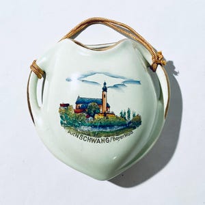 Könnte beinhalten: Eine herzförmige, cremefarbene Keramik-Wandtasche mit einer gemalten Landschaftsszene einer Kirche und dem Text "ARNSCHWANG/BayernWald". Sie hat einen goldfarbenen Rand und einen goldfarbenen Seilgriff.
