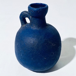 Może przedstawiać: Mały, niebieski ceramiczny wazon o zaokrąglonym korpusie i pojedynczym uchwycie. Wazon ma fakturowaną powierzchnię i lekko rozszerzający się otwór.