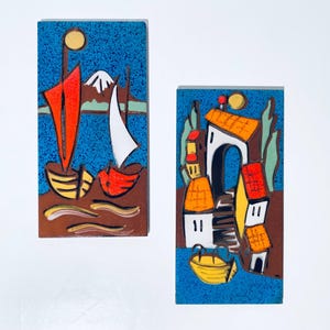 Könnte beinhalten: Zwei rechteckige Keramikfliesen mit farbenfrohen Küstenszenen. Eine Fliese zeigt Boote mit roten und weißen Segeln, die andere Gebäude mit orangefarbenen Dächern und einem gelben Boot vor blauem Hintergrund.