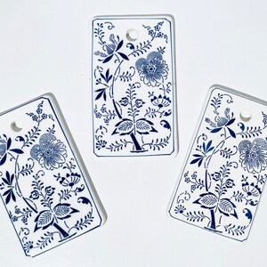 Könnte beinhalten: Drei rechteckige Fliesen aus weißer Keramik mit blauen Blumendesigns. Jede Fliese hat oben ein Loch zum Aufhängen.