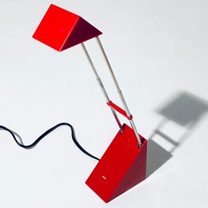 Könnte beinhalten: Eine rote Schreibtischlampe mit einem dreieckigen Schirm und einem verstellbaren Arm aus Chrom. Die Lampe hat ein schwarzes Kabel.