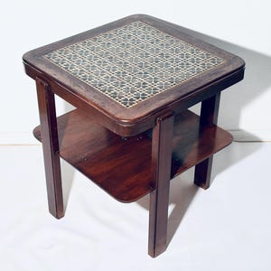 May include: A square, dark wood side table with a patterned tile top. The table has a lower shelf and four straight legs. The tile design features a repeating pattern of blue and white shapes. The table is approximately 20 inches tall.
