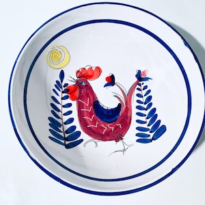 Puede incluir: Un cuenco de cerámica blanca con un borde azul y un diseño pintado a mano de un gallo con detalles rojos, azules y amarillos. El gallo está rodeado de hojas estilizadas azules y blancas.