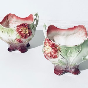 Könnte beinhalten: Zwei antike Keramik-Blumentöpfe mit verzierten Blumendesigns. Die Töpfe sind weiß mit grünen und roten Akzenten und haben erhabene Blumendetails.
