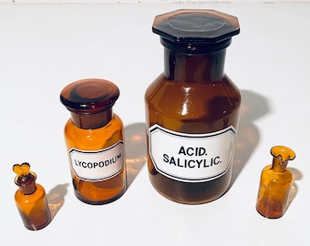 May include: Four amber glass bottles with white labels. The largest bottle has a label that reads "ACID. SALICYLIC.". The next largest bottle has a label that reads "LYCOPODIUM". The two smallest bottles have labels that are too small to read.