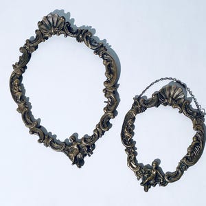 Könnte beinhalten: Zwei verzierte goldfarbene Bilderrahmen aus Metall mit aufwendigen Designs. Der größere Rahmen ist oval und der kleinere Rahmen ist rund.