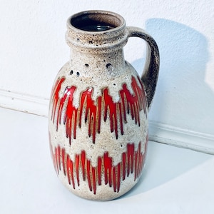 Könnte beinhalten: Eine weiße Keramikvase mit einem roten und braunen Tropf-Glazurmuster. Die Vase hat einen Henkel und einen runden Körper.