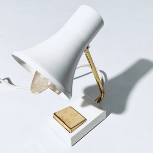 Könnte beinhalten: Eine weiße Tischlampe mit einem goldenen Metallarm und Sockel. Die Lampe hat einen weißen Schirm und eine klare Glühbirne. Die Lampe hat einen goldenen Kippschalter am Sockel.