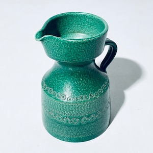 Puede incluir: Una jarra de cerámica verde con una superficie texturizada y un diseño decorativo alrededor del centro. La jarra tiene una boca ancha y un asa.