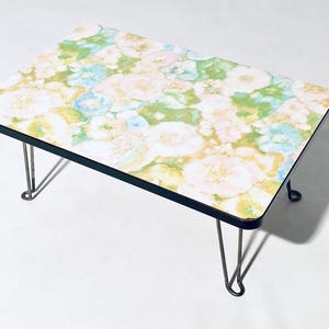 Op de afbeelding: Een kleine rechthoekige tafel met een bloemenpatroon. Het tafelblad is versierd met witte, gele, groene en blauwe bloemen. Het heeft een zwarte rand en dunne metalen haarspeldpoten. De tafel is ongeveer 60 cm lang.