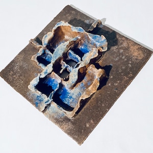 Könnte beinhalten: Eine quadratische Wandskulptur aus Keramik mit einem strukturierten, abstrakten Design. Die Skulptur ist aus braunem und blauem Ton gefertigt und hat ein erhabenes, dreidimensionales Design.