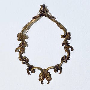 Könnte beinhalten: Ein verzierter Metallrahmen in Goldfarbe mit einem dekorativen Design. Der Rahmen ist tropfenförmig und hat ein zartes, filigranes Muster.