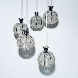 Op de afbeelding: Vijf hangende decoratieve ornamenten in de vorm van granaatappels. Elk ornament is gemaakt van heldere, facetgeslepen kralen en heeft een goudkleurig metalen frame en een donkere, gestileerde kroon. De ornamenten hangen aan witte koorden tegen een witte achtergrond.