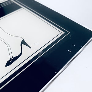 May include: A black and white framed print of a woman's legs in high heels. The print is framed with a black and white border.