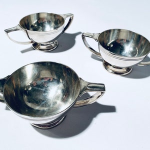 May include: Three silver cups with handles, each with a round base and a smooth, reflective surface.
