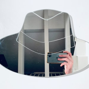 Puede incluir: Un espejo con marco plateado con forma de sombrero, reflejando a una persona sosteniendo un teléfono inteligente.