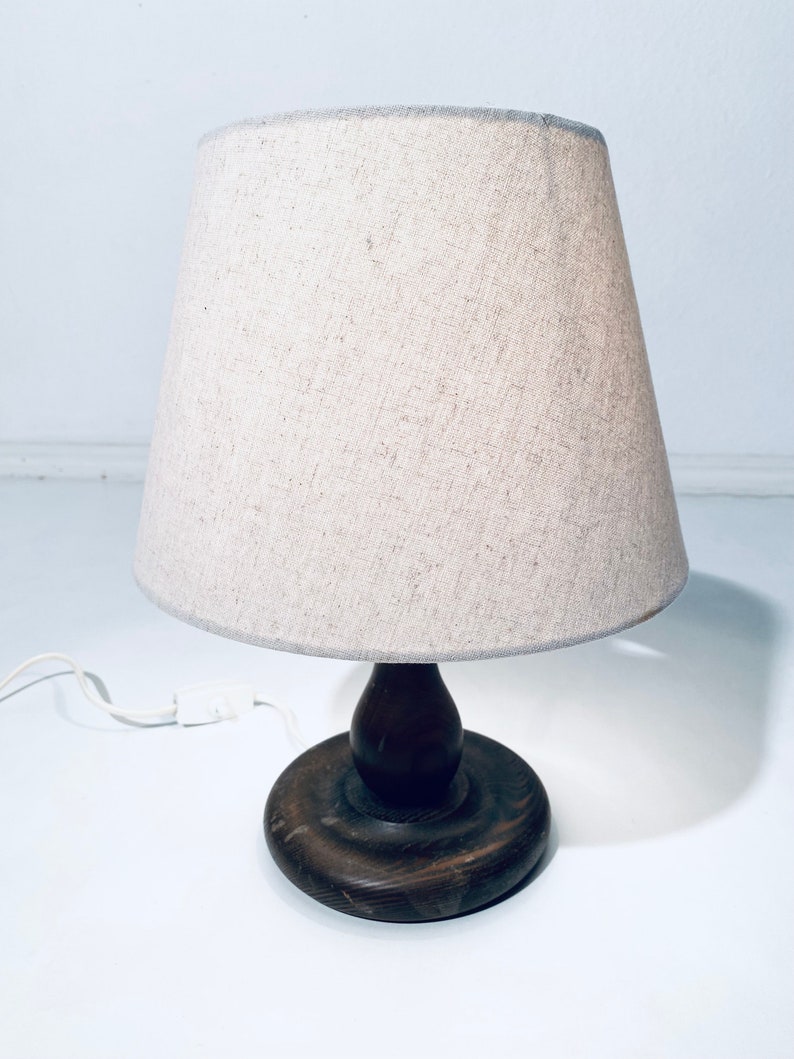 May include: A wooden table lamp with a beige linen lampshade. The lamp has a round wooden base and a wooden stem.