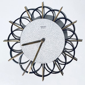 May include: A white clock face with black crackle glaze and gold hour and minute hands. The clock face is surrounded by a black metal frame with gold accents.