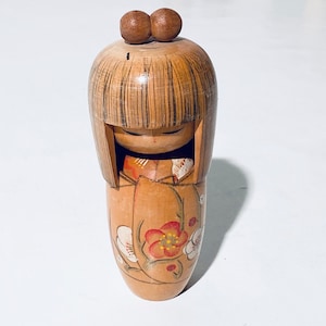 Könnte beinhalten: Eine traditionelle japanische Holzpuppe mit einem bemalten Blumendesign auf ihrem Kimono. Die Puppe hat eine braune Holzfrisur und geschlossene Augen.