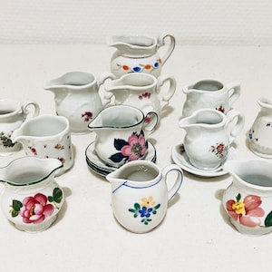 May include: A collection of ten small, white porcelain creamer pitchers with floral designs. The pitchers are arranged in a circle on a white surface.