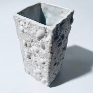 Könnte beinhalten: Eine weiße Keramikvase mit einer strukturierten Oberfläche und einer quadratischen Form. Die Vase ist mit kleinen, erhabenen Muscheln verziert.