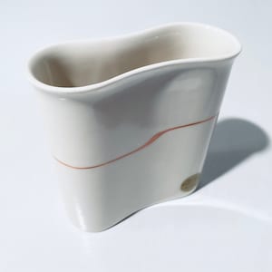 以下が含まれることがあります： 波打つような非対称な形をした白い陶器の花瓶。花瓶の中央に細いオレンジ色の線が引かれている。