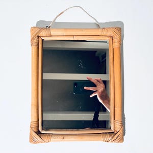 Könnte beinhalten: Rechteckiger Spiegel mit Bambusrahmen. Der Rahmen hat ein geflochtenes Design an den Ecken. Der Spiegel hat eine hellbraune Farbe und eine weiße Schnur zum Aufhängen. Die Reflexion zeigt eine Person, die ein Telefon hält.