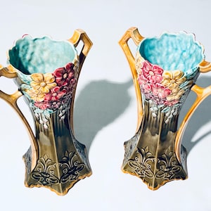 May include: Two matching ceramic vases with a floral design. The vases have a light blue interior, with yellow, pink, and red flowers. The exterior is a dark green with gold accents and decorative patterns. The vases have handles.