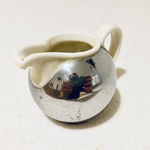 Könnte beinhalten: Ein kleiner, silberner Milchkännchen mit einem weißen, gebogenen Ausguss und einem Griff. Der Milchkännchen ist mit einer hellfarbigen Flüssigkeit gefüllt.