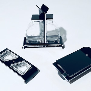 May include: A black and white condiment set with three pieces. The set includes a two-bottle holder, a small dish with two compartments, and a single dish.