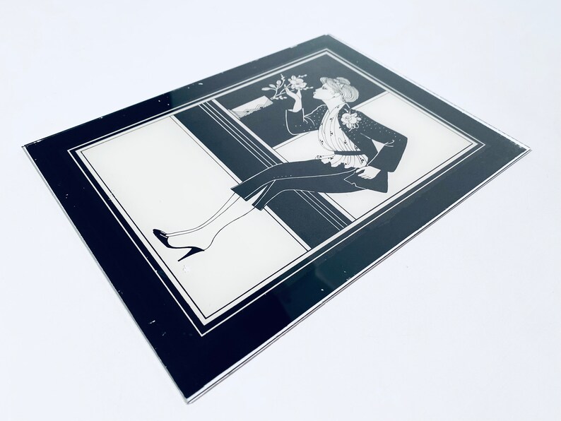 May include: Black and white framed illustration of a woman in a stylish outfit, sitting on a ledge and holding a flower.