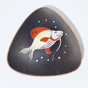 Pode incluir: Um prato de cerâmica triangular com fundo preto e um peixe branco com detalhes vermelhos e laranja. O peixe tem um corpo branco com escamas brancas e pretas, uma boca vermelha e uma cauda vermelha e laranja. O prato é decorado com pequenos pontos azuis.