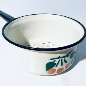 Könnte beinhalten: Weißer Email-Sieb mit schwarzem Rand und einem grünen und orangefarbenen Fruchtmuster. Das Sieb hat einen schwarzen Griff.