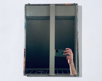 May include: A silver framed mirror with a reflection of a person taking a selfie with a black smartphone.